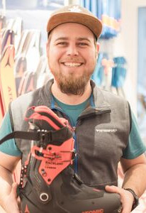 Lächelnder Mann in einem Skigeschäft, der einen schwarz-roten Skischuh hält, umgeben von Skiausrüstung.