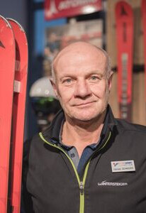 Lächelnder Mann in einem Intersport-Skigeschäft, stehend neben roten Atomic-Skiern und Wintersportausrüstung.