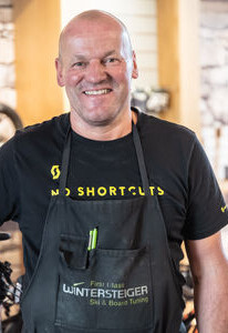 Lächelnder Techniker in einer WINTERSTEIGER Ski- und Board-Service-Schürze, stehend in einer Fahrradwerkstatt.