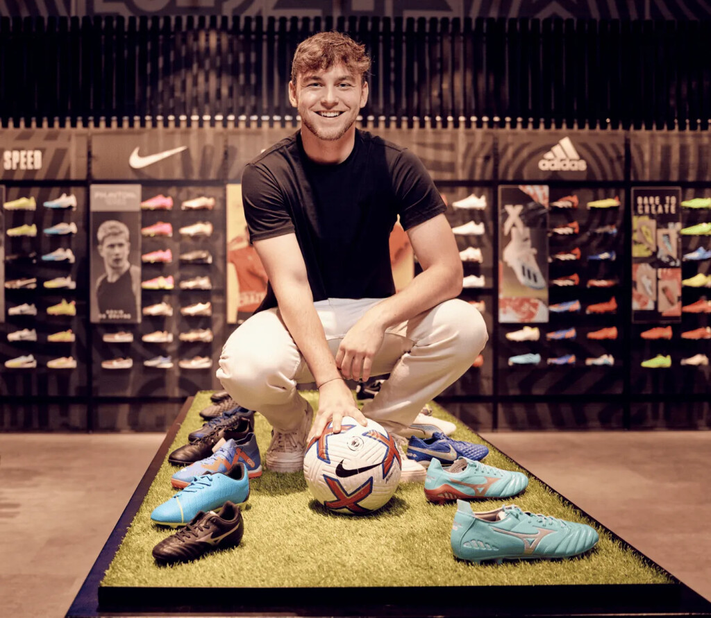 Ein lächelnder Mann hockt mit einem Fußball und Stollenschuhen, die in einem modernen Fußball-Einzelhandelsgeschäft ausgestellt sind.
