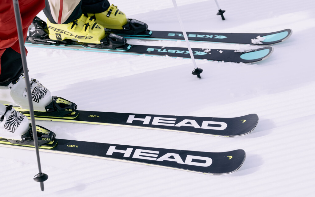Nahaufnahme von HEAD- und KÄSTLE-Skiern mit Skischuhen und Skistöcken auf einer präparierten Schneepiste.
