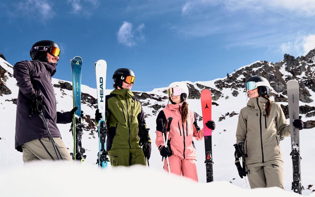 Vier Skifahrer in bunter Skibekleidung und Helmen, die Skier halten, auf einem sonnigen, verschneiten Berg.