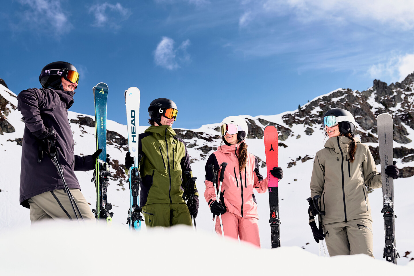 Vier Skifahrer in farbenfroher Skibekleidung und Helmen, die Skier halten, auf einem sonnigen, verschneiten Berg.