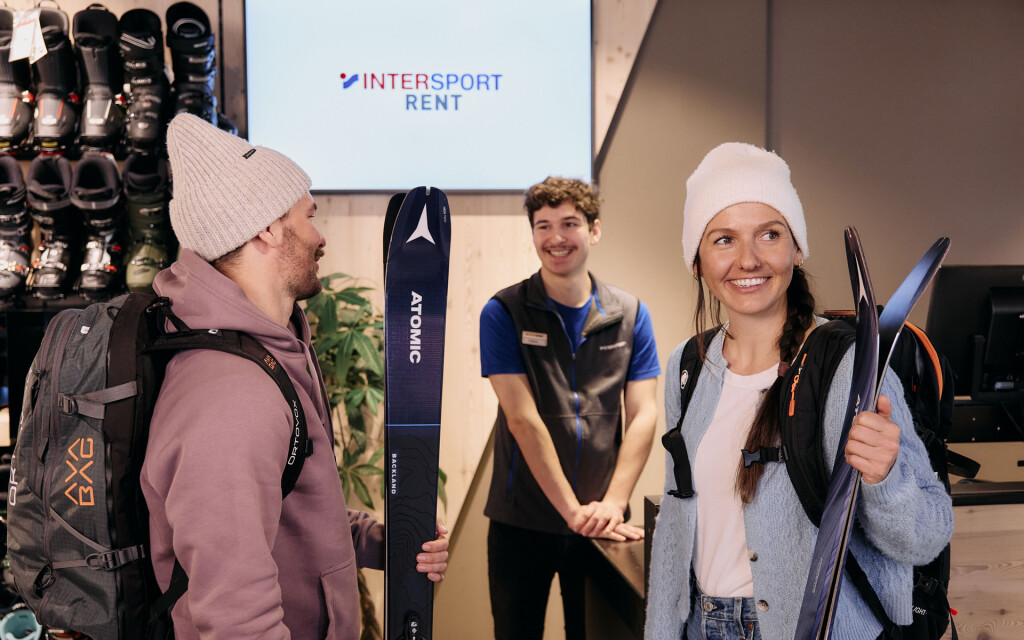 Zwei Kunden mit Atomic-Skiern interagieren mit Mitarbeitern von Intersport Rent in einem Skiausrüstungsgeschäft.