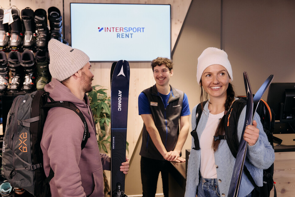Zwei Kunden mit Atomic-Skiern interagieren mit Mitarbeitern von Intersport Rent in einem Skiausrüstungsgeschäft.