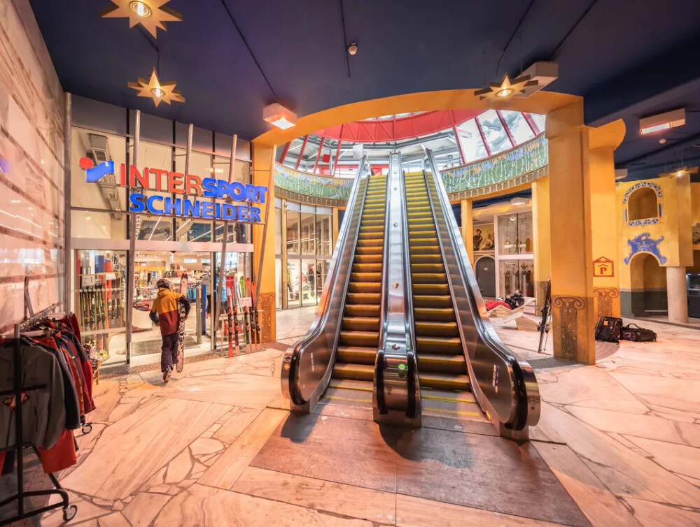 Innenansicht eines modernen Einkaufszentrums mit einer großen Rolltreppe, einem Intersport-Geschäft und einem Kunden.