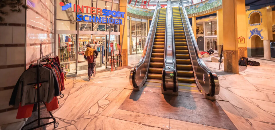 Innenansicht eines Einkaufszentrums mit einer markanten Rolltreppe, dem Intersport-Ladeneingang und Bekleidungsauslagen.
