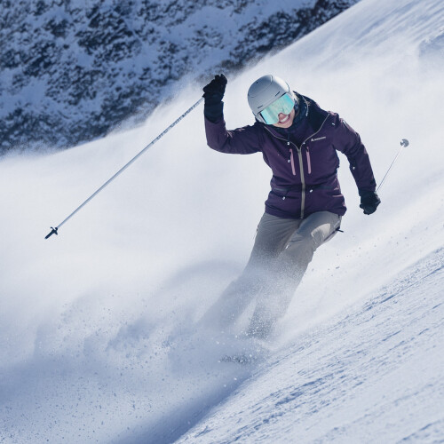 Ein Skifahrer in lila Jacke und Helm carvt einen verschneiten Berghang hinunter und wirbelt frischen Pulverschnee auf.