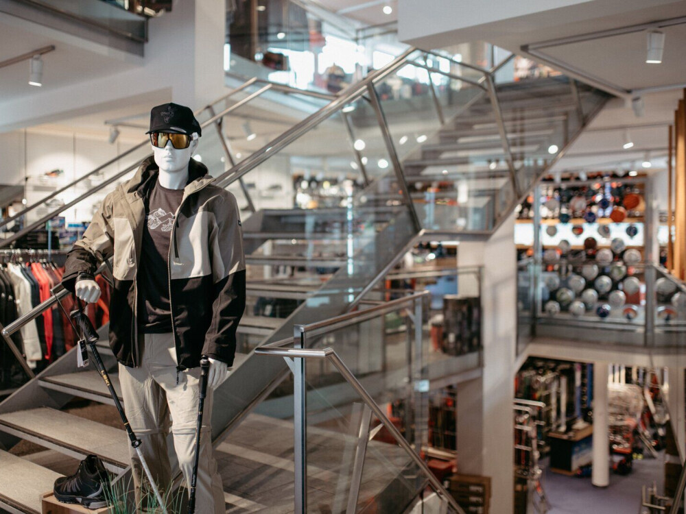 Sportbekleidungs-Schaufensterpuppe mit Jacke, Mütze, Maske, Sonnenbrille und Trekkingstock auf einer Treppe in einem modernen Einzelhandelsgeschäft.