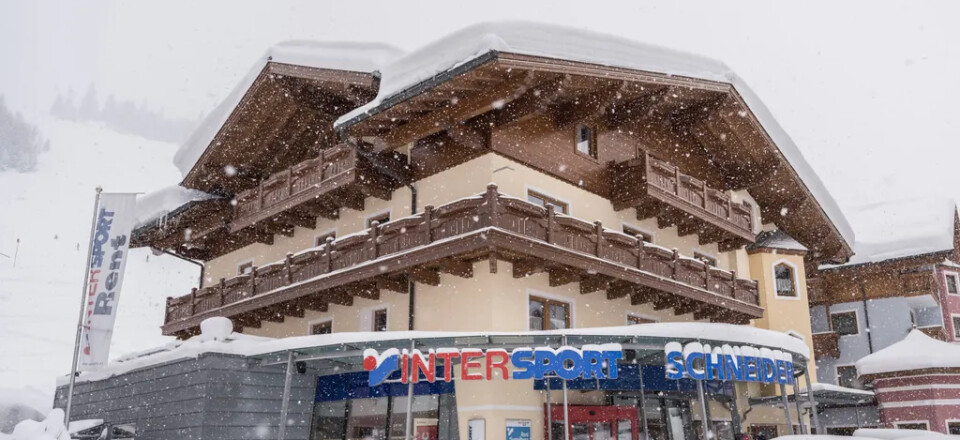 Außenansicht eines InterSport Schneider Wintersportgeschäfts in einem verschneiten Alpenort mit geparkten Autos und starkem Schneefall.