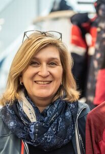Lächelnde Frau mit Brille und Schal, die eine rote Steppjacke in einem Outdoor-Bekleidungsgeschäft hält.