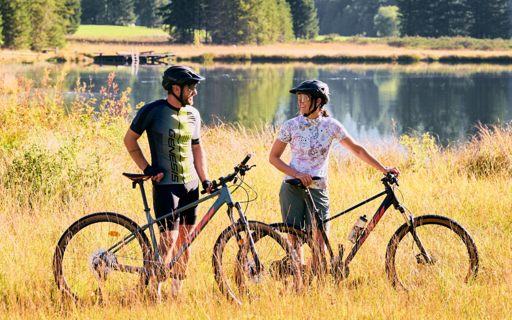 Ein Paar mit Mountainbikes genießt die malerische Aussicht auf einen Bergsee mit Wäldern und Gipfeln.
