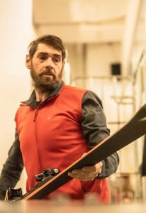 Bärtiger Mann in roter Weste hält einen schwarzen Ski und steht in einem Indoor-Skiausrüstungsbereich.