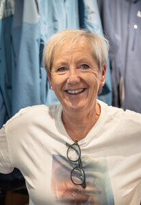 Lächelnde reife Frau mit kurzen blonden Haaren, weißem T-Shirt und Brille in einem Bekleidungsgeschäft.