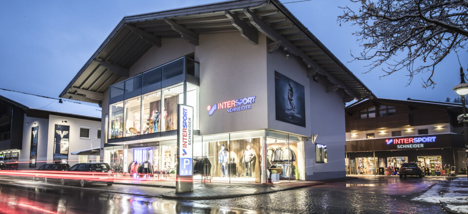 Sportgeschäft Intersport mit beleuchteten Fenstern auf nasser Straße in der Dämmerung, Lichtspuren, Alpenstadt.