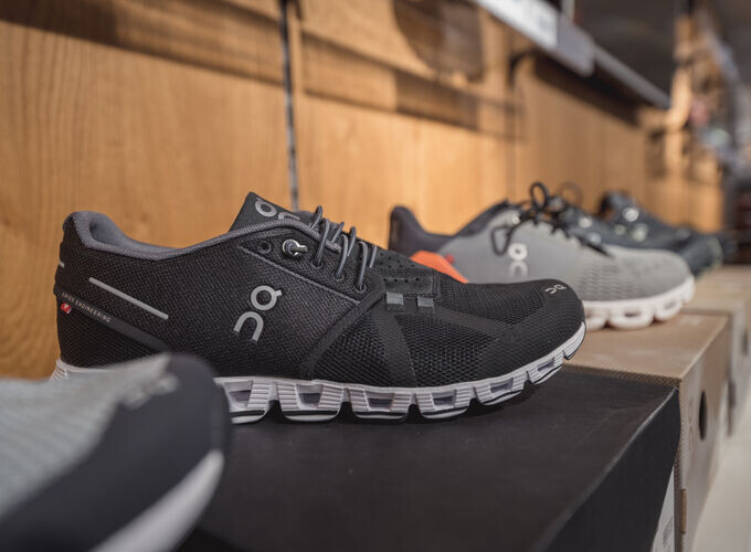 Schwarze On Cloud Laufschuhe auf einem Ausstellungsregal in einem Einzelhandelsgeschäft, mit grauen Sportschuhen im Hintergrund.