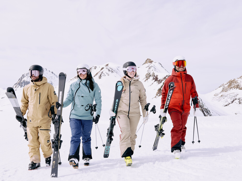Vier Skifahrer in kompletter Winterausrüstung gehen durch eine verschneite Berglandschaft und tragen Skier.