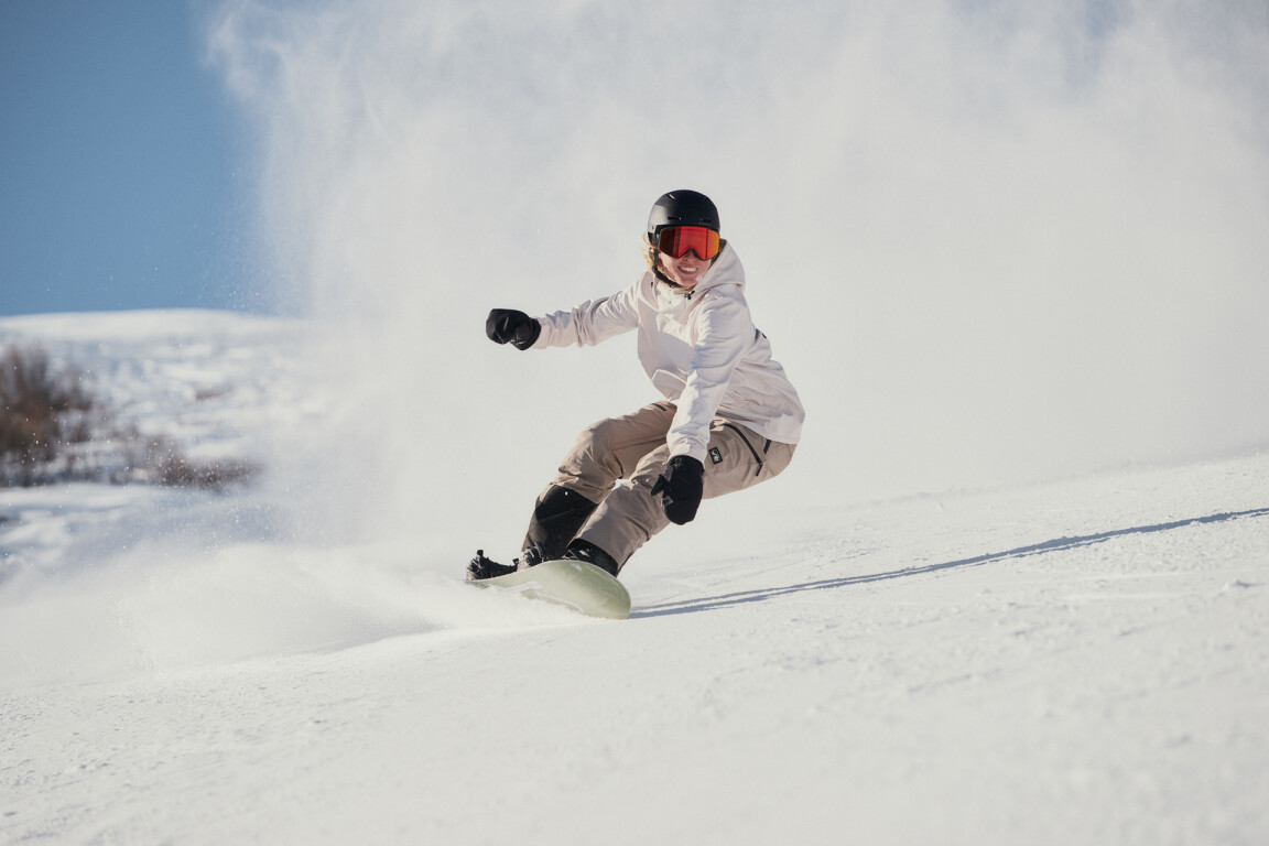 Eine weibliche Snowboarderin carvt einen dynamischen Turn auf einem verschneiten Berghang und wirbelt frischen Pulverschnee auf.