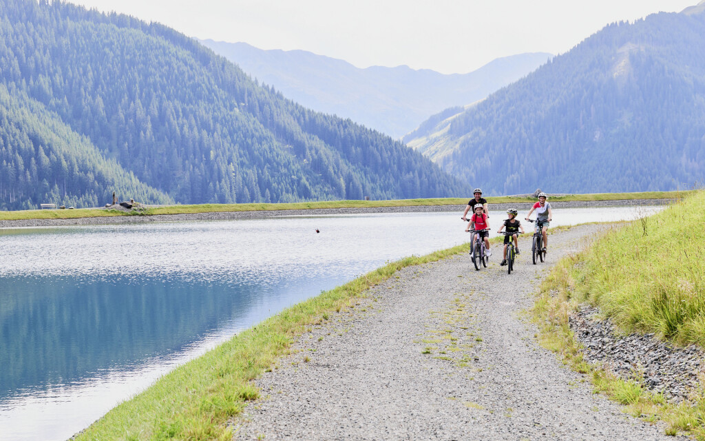 Eine vierköpfige Familie radelt auf einem Schotterweg neben einem Bergstausee, umgeben von grünen Gipfeln.