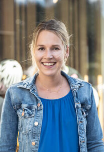 Lächelnde junge blonde Frau in einer Jeansjacke, lässig vor einem Einzelhandelsgeschäft stehend.