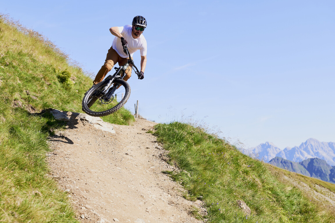 A mountain biker in the air on a steep dirt path with green hills and distant mountain peaks.