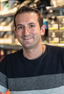 Lächelnder Mann in einem gestreiften T-Shirt, stehend in einem Gang mit verpackten Produkten in Regalen.
