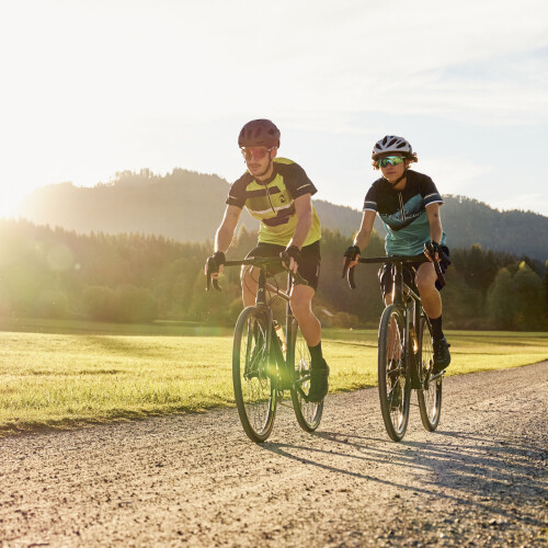 Zwei Radfahrer auf Gravelbikes fahren einen sonnigen Schotterweg durch ein grasbewachsenes Feld mit Bergen bei Sonnenuntergang.