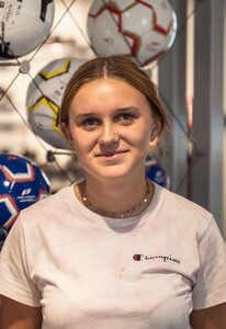 Lächelnde junge Frau in einem Champion T-Shirt, steht in einem Sportgeschäft mit ausgestellten Fußbällen.