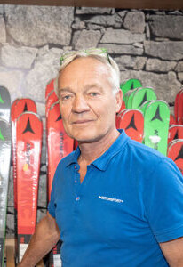 Mann in blauem Poloshirt in einem Skigeschäft, stehend vor Reihen von bunten Atomic-Skiern.