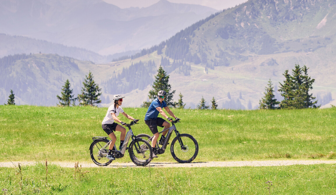 Two people are riding e-bikes on a picturesque outdoor path through a green mountain landscape.
