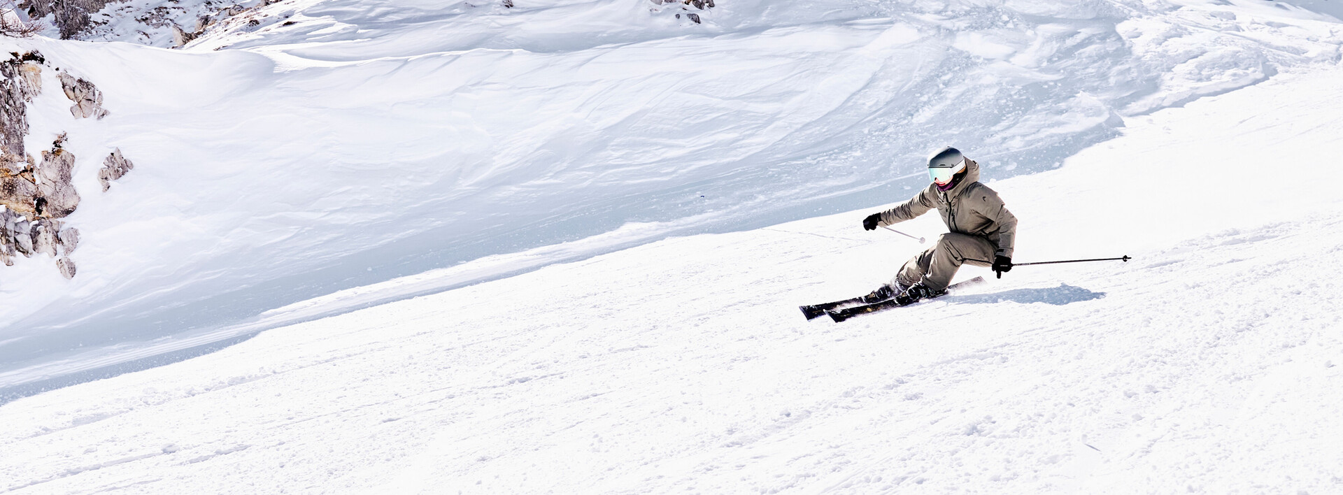 Ein Skifahrer mit Helm und Skibrille fährt einen weiten, schneebedeckten Berghang unter klarem Himmel hinab.