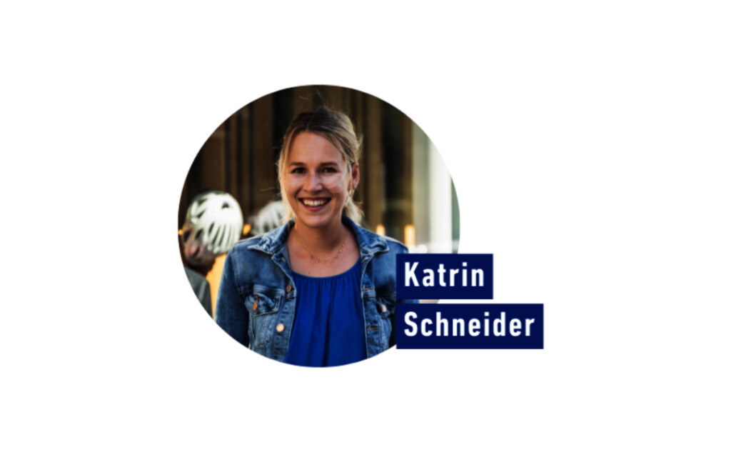 Professionelles Porträt der lächelnden Katrin Schneider in Jeansjacke und blauem Oberteil, mit Namenseinblendung.