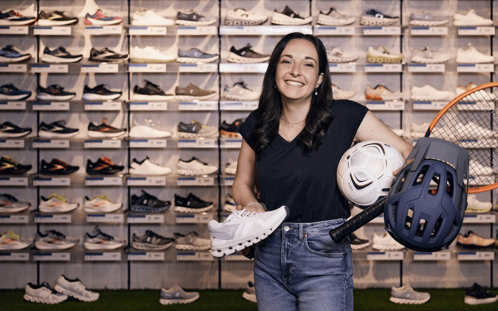 Smiling woman holding a running shoe, soccer ball, helmet, and tennis racket in a running shoe store.