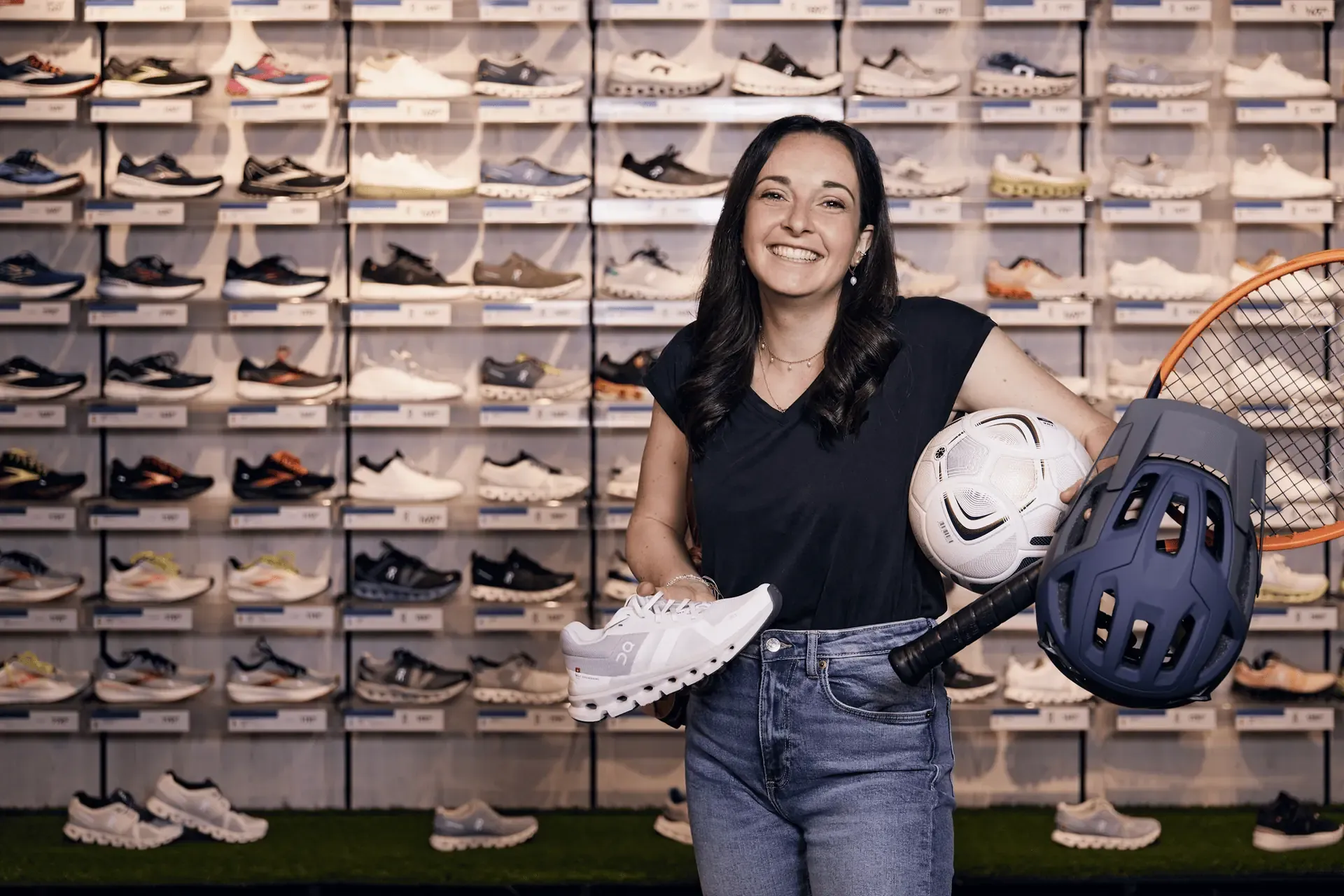 Eine lächelnde Frau hält einen weißen Laufschuh, einen Fußball, einen Helm und einen Tennisschläger in einem Sportschuhgeschäft.