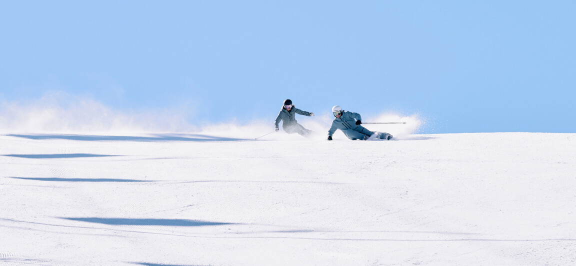 Sonnige Bergskipiste mit zwei Skifahrern: einer carvt, der andere stürzt und wirbelt dabei Schneespritzer auf.