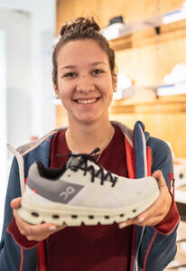 Lächelnde Frau, die einen weiß-grauen Laufschuh hält und dabei Sportschuhe in einem Einzelhandelsgeschäft präsentiert.